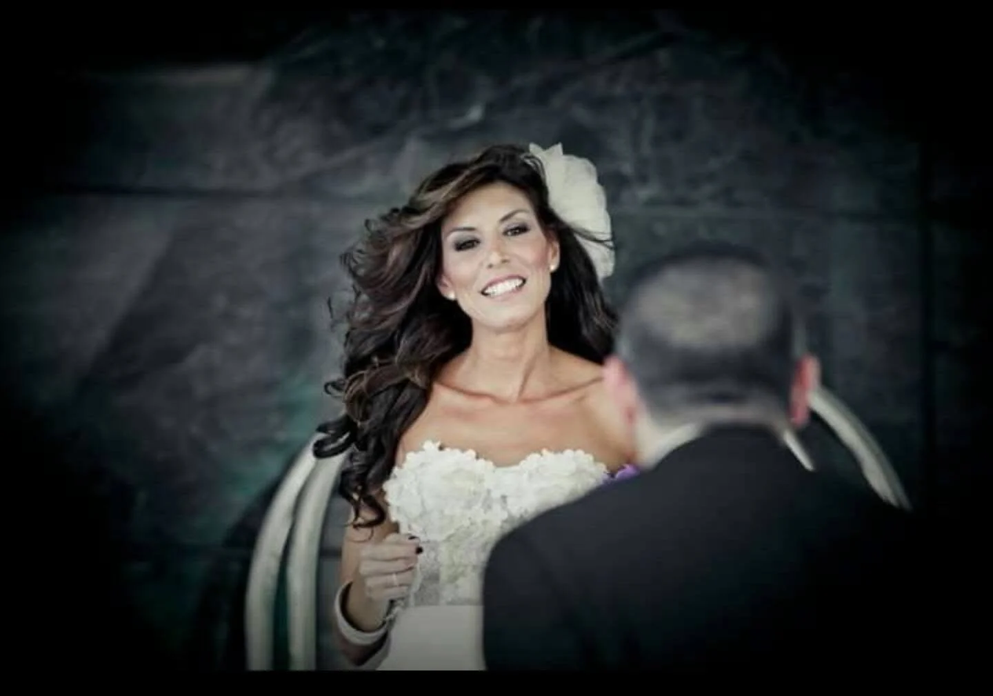 A bride with long, dark, wavy hair smiling at her groom, holding a white bouquet, during their wedding ceremony.