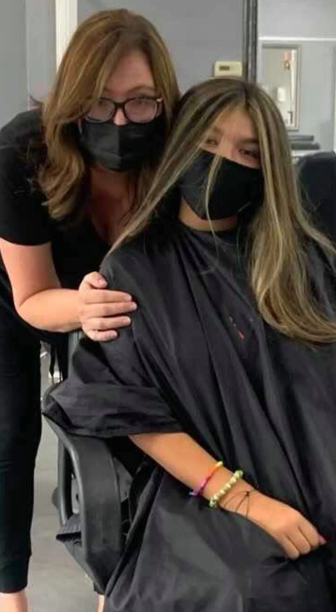 Two women, wearing black masks, one with glasses and one with long blonde hair, are posing in a hair salon. The woman with long blonde hair is seated in a salon chair with her hair partially covered with a black cape. The woman with glasses is standi