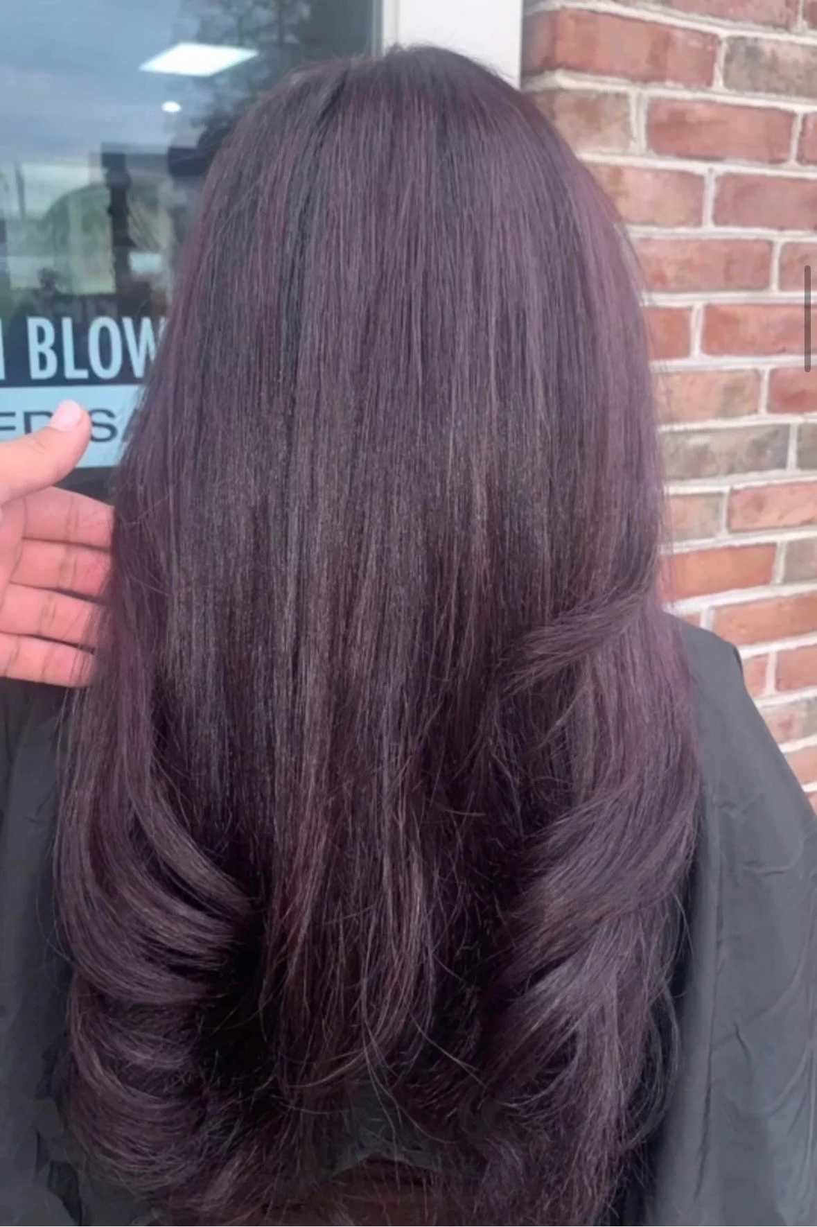 Back of a woman's head with long, straight, and shiny purple hair styled with gentle curls at the ends, standing outdoors in front of a brick wall and a window.