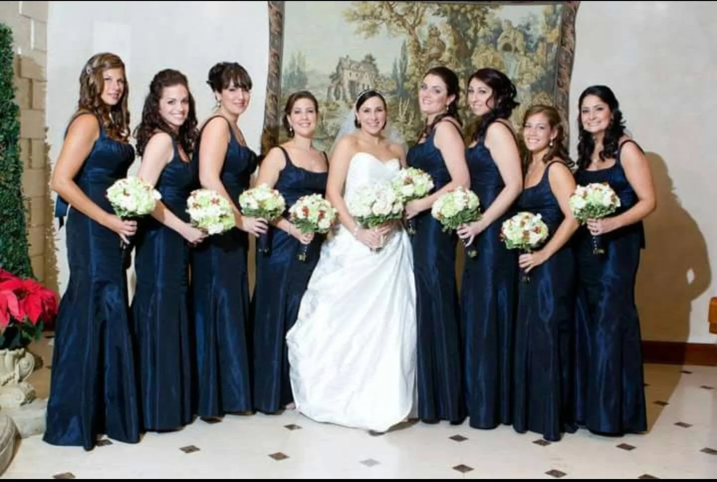 A bride in a white wedding dress standing with her bridesmaids in matching dark blue dresses, each holding a bouquet of white and pink flowers, in an indoor setting with a tapestry and tiled floor.