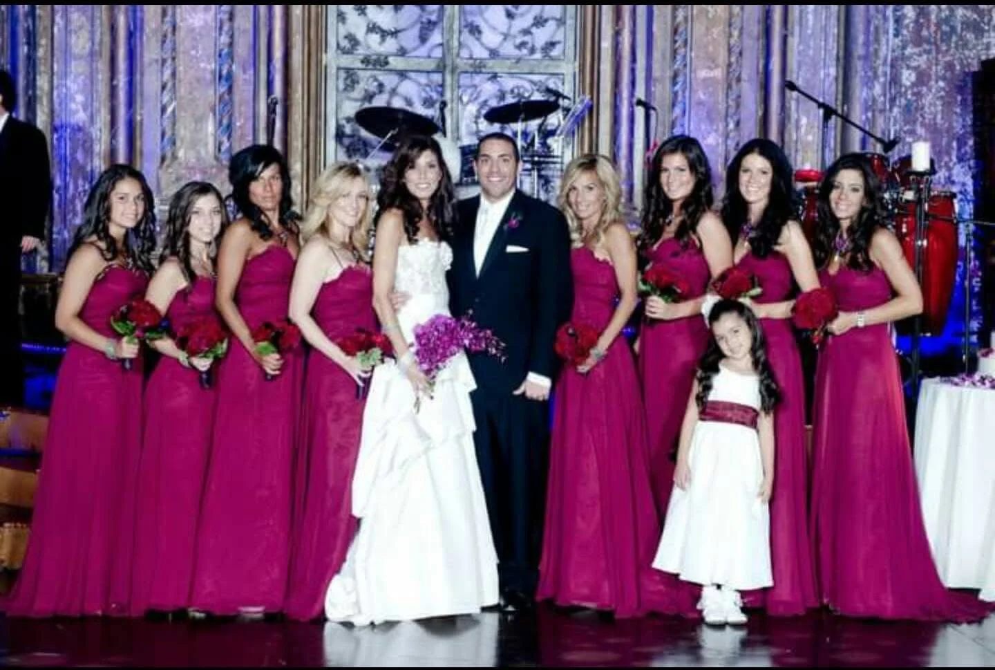 A wedding party with the bride, groom, ten bridesmaids, and a young flower girl, all dressed in formal attire, standing in a decorated venue with musical instruments in the background.