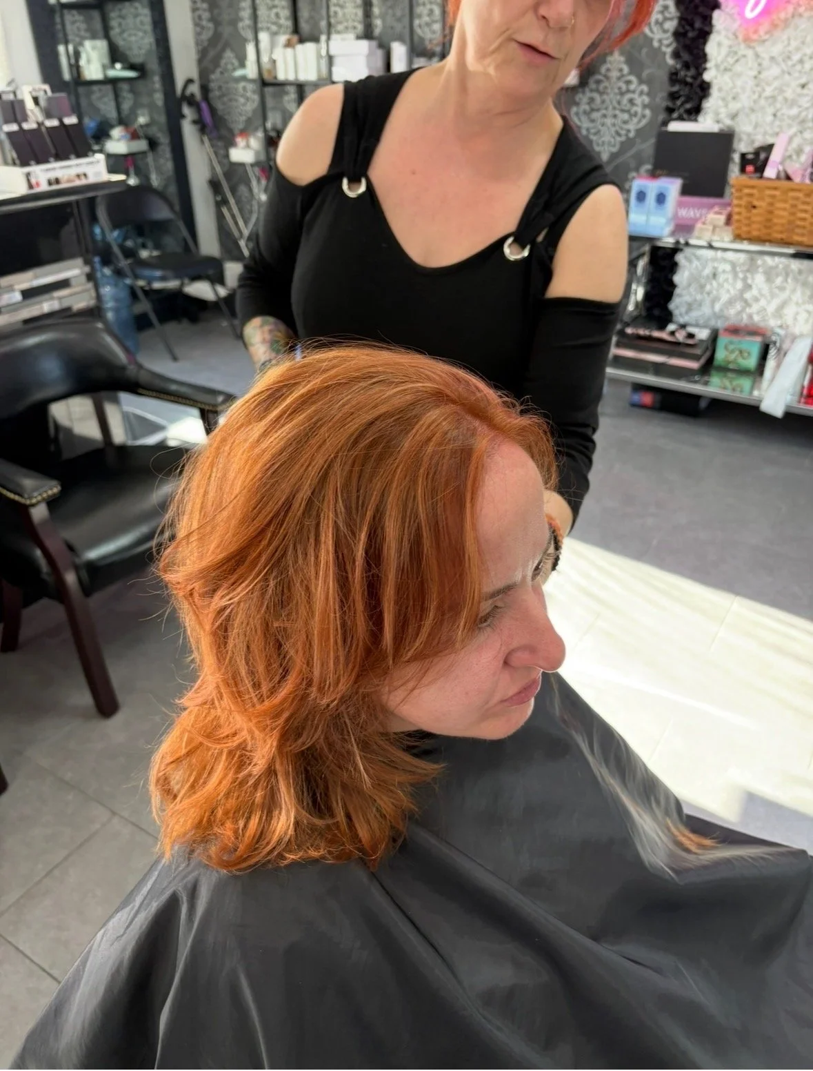 A woman with shoulder-length red hair sitting in a salon chair, getting her hair styled by a hairstylist.