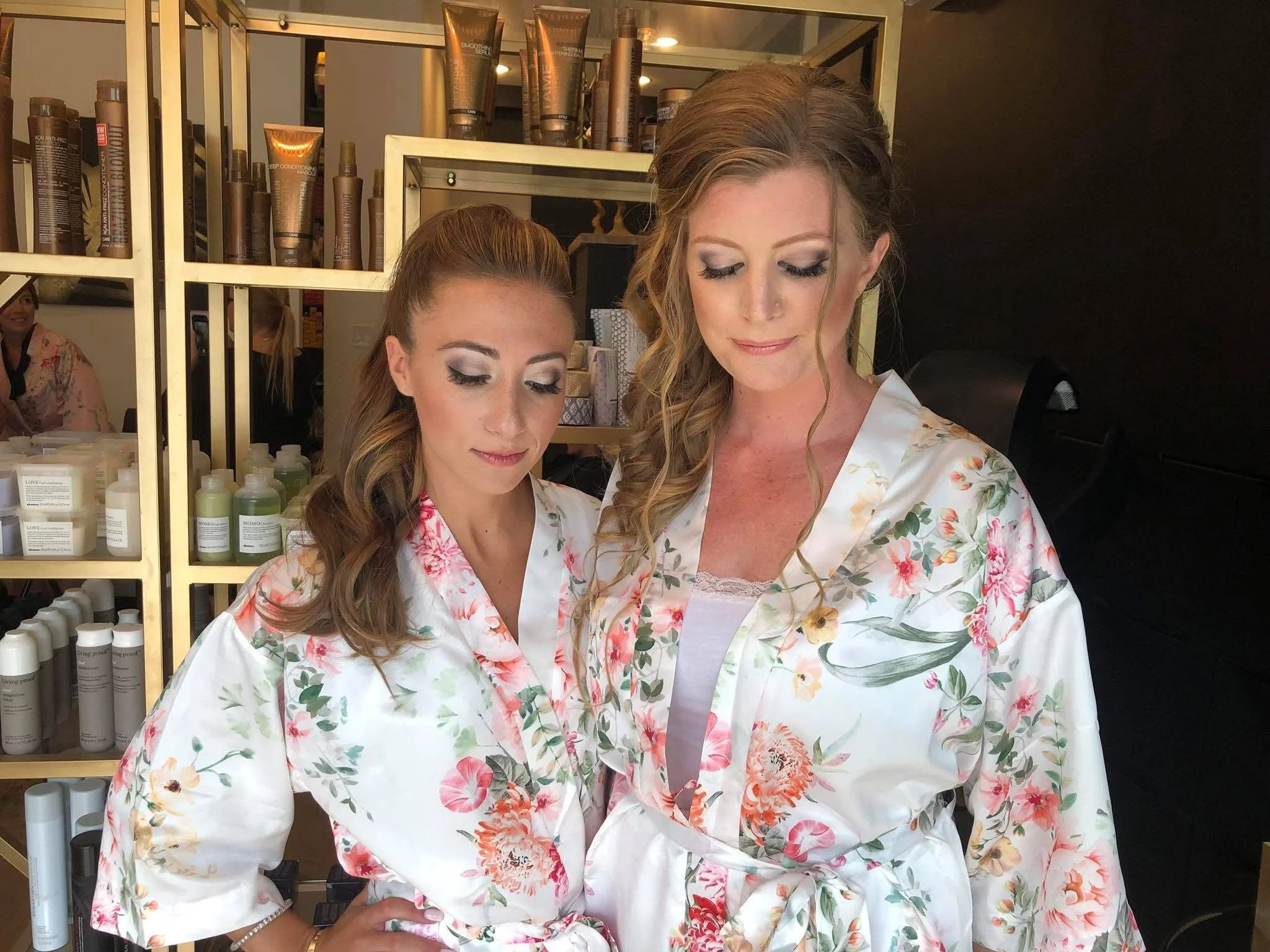 Two women in floral robes with makeup and hairstyles, standing in front of shelves with hair and beauty products, preparing for a special event or photo shoot.