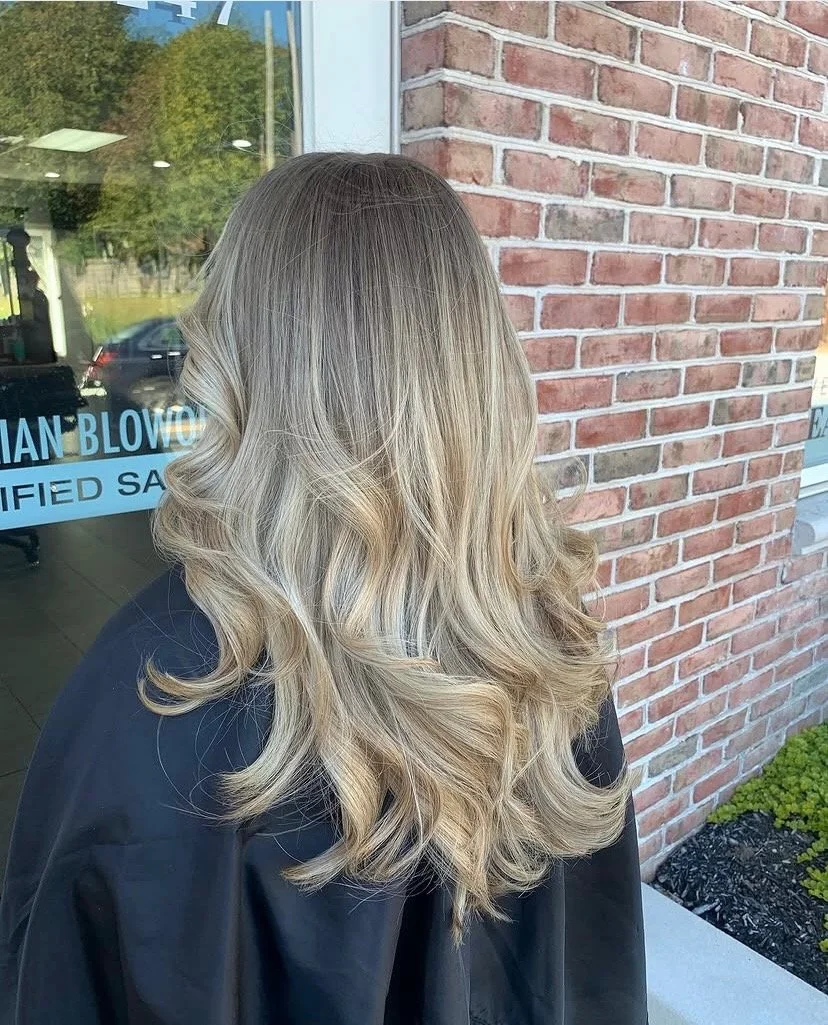 Back view of woman with long, blonde, wavy hair outside a building with a brick wall and window.