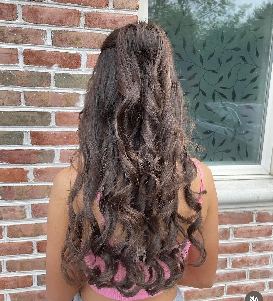 Back view of a woman with long, wavy brown hair standing outside near a brick wall and window.