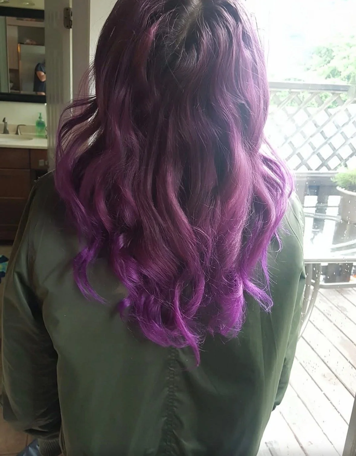 Person with wavy hair dyed in shades of purple, standing indoors near a window with a view of a deck and outdoor furniture.