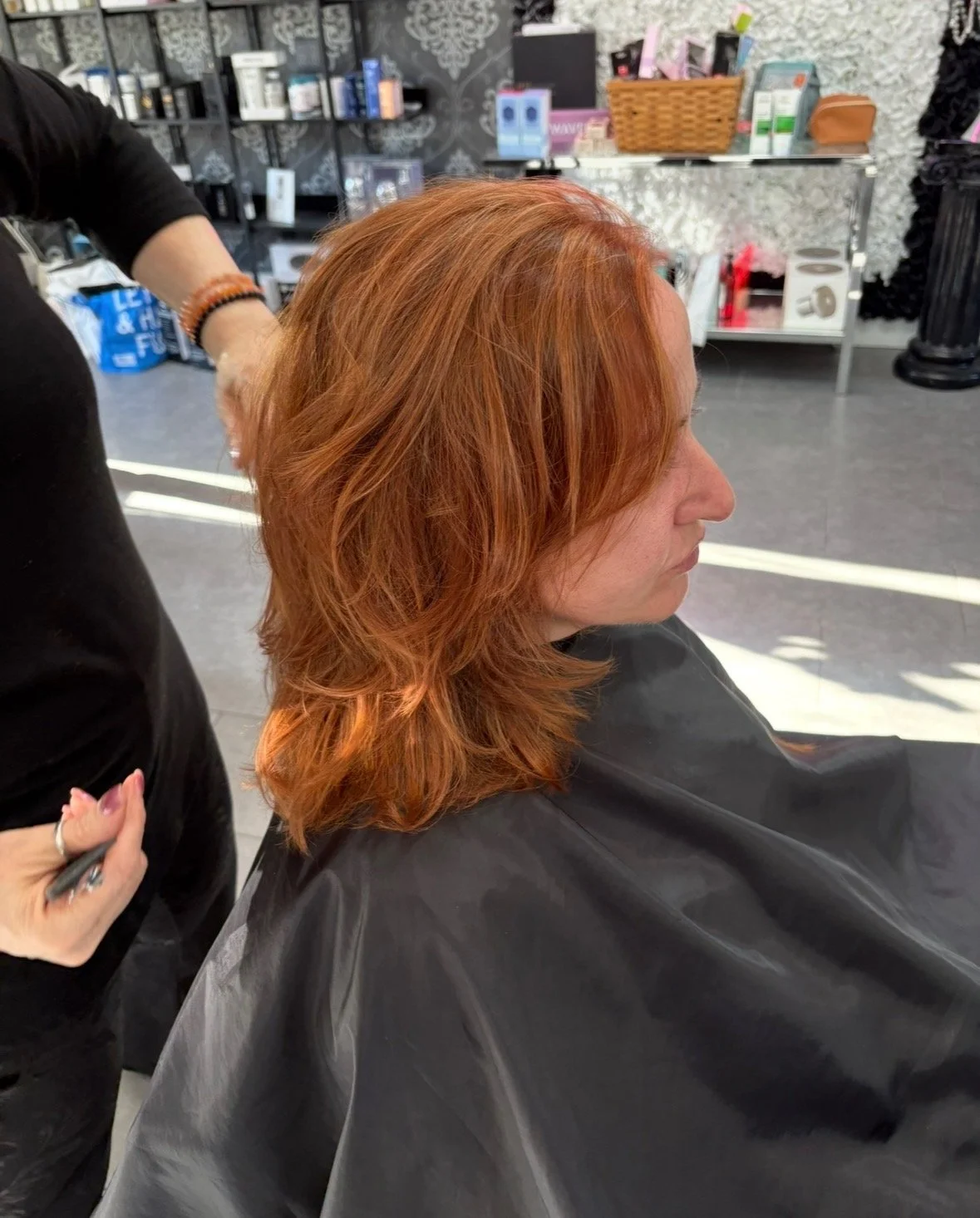 Woman with shoulder-length wavy auburn hair sitting in a salon chair with a black cape, while a hairstylist works on her hair. The salon background has shelves with hair and beauty products.