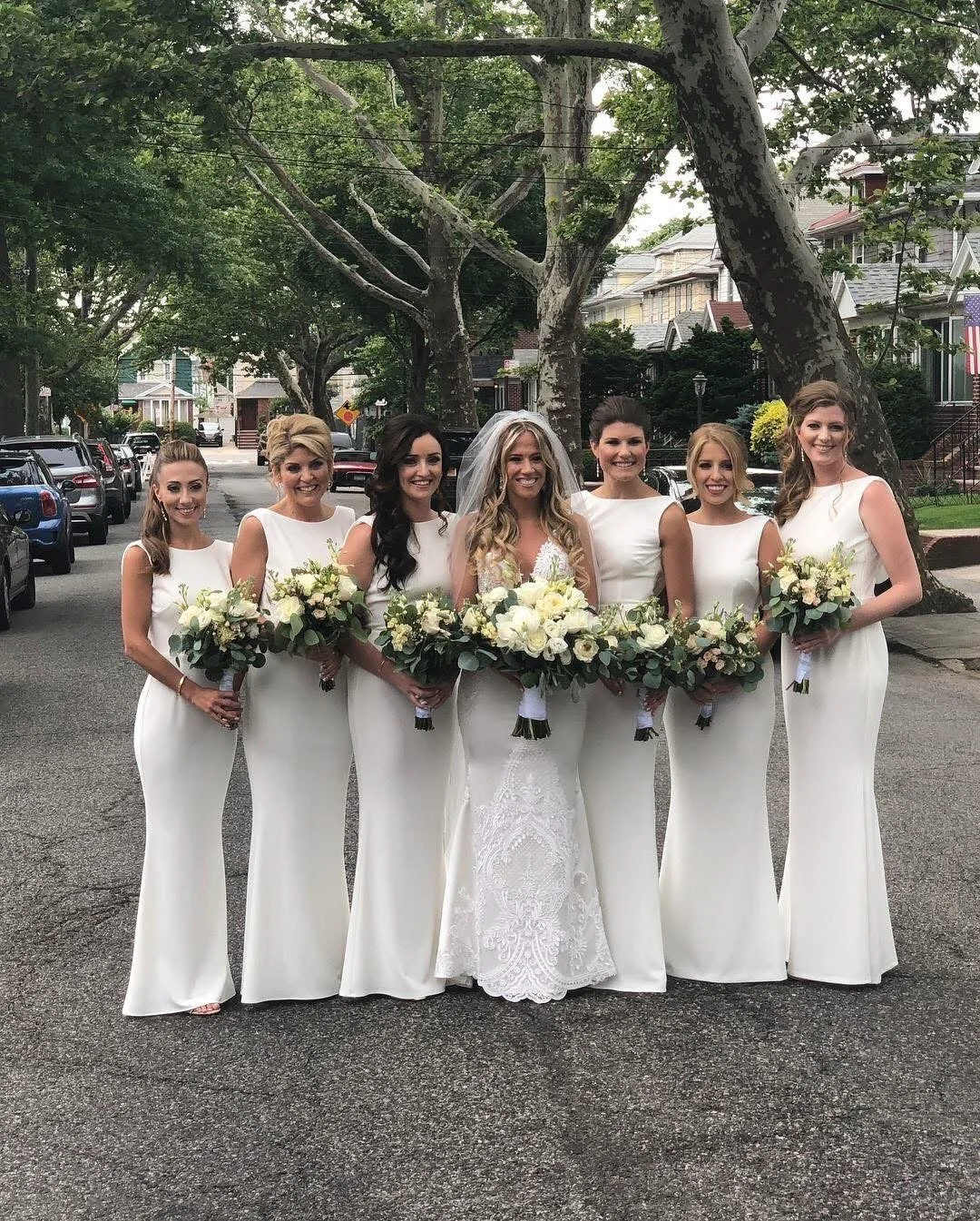 Seven women dressed in white wedding and bridesmaid gowns, holding bouquets of white flowers, standing outdoors on a residential street with trees and houses in the background.