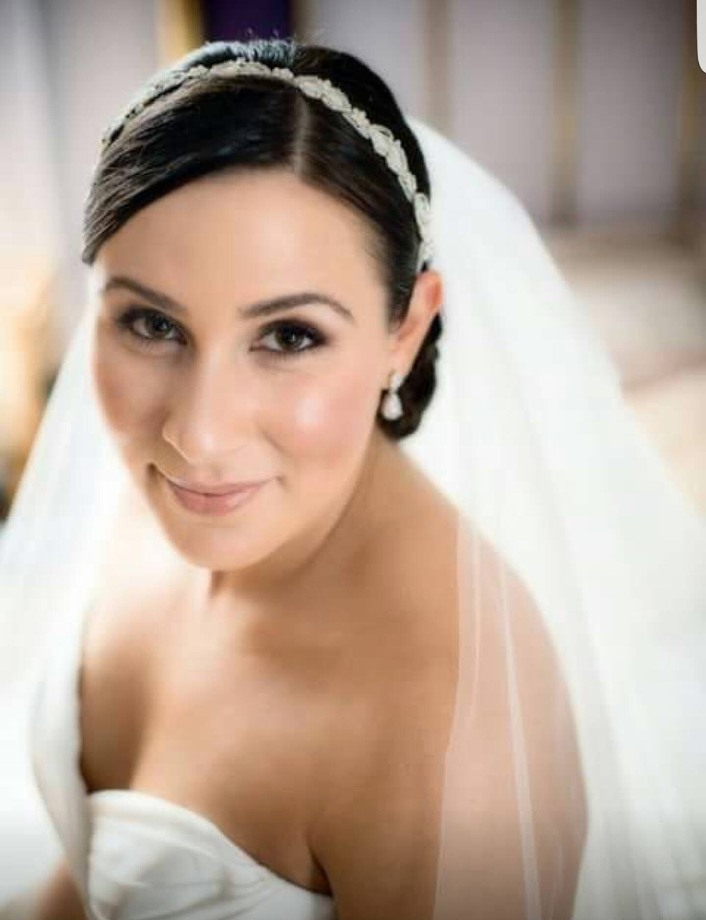 Close-up of a smiling woman with dark hair, wearing a pearl headband and pearl earrings, partially wrapped in white fabric, indoors.