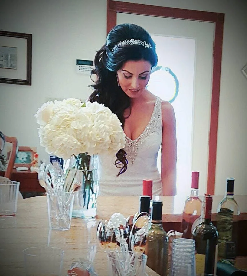 A bride with elaborate dark hair, a silver tiara, and a white wedding gown with lace details, standing at a table with a large bouquet of white flowers, surrounded by bottles of wine, and glasses.