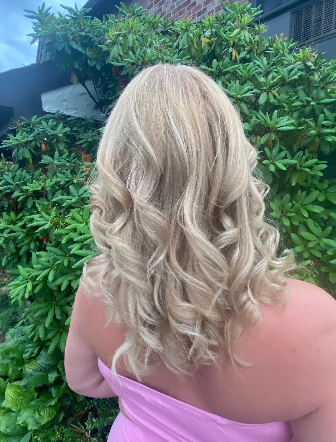A woman with long, blonde, curly hair wearing a pink strapless dress, standing outdoors with green leafy bushes in the background.