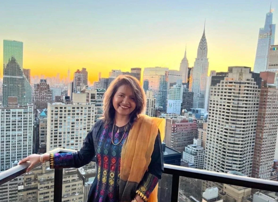 A woman smiling and standing on a balcony with a city skyline at sunset in the background, wearing a colorful traditional Indian outfit and a yellow dupatta.