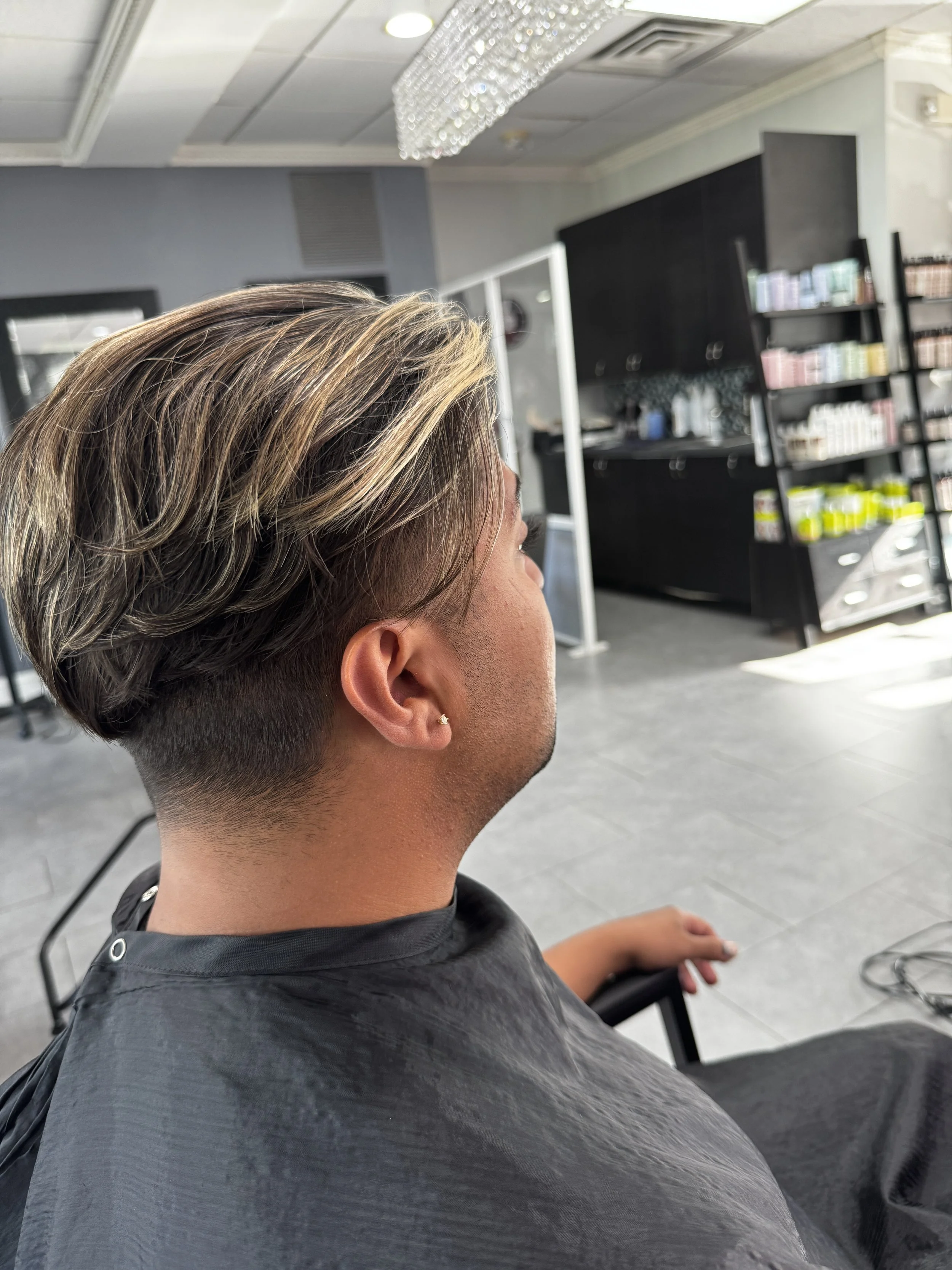 A person with a short undercut haircut and blond highlights is sitting in a salon chair at a hair salon, wearing a black cape. The salon has shelves with hair products and a large mirror.