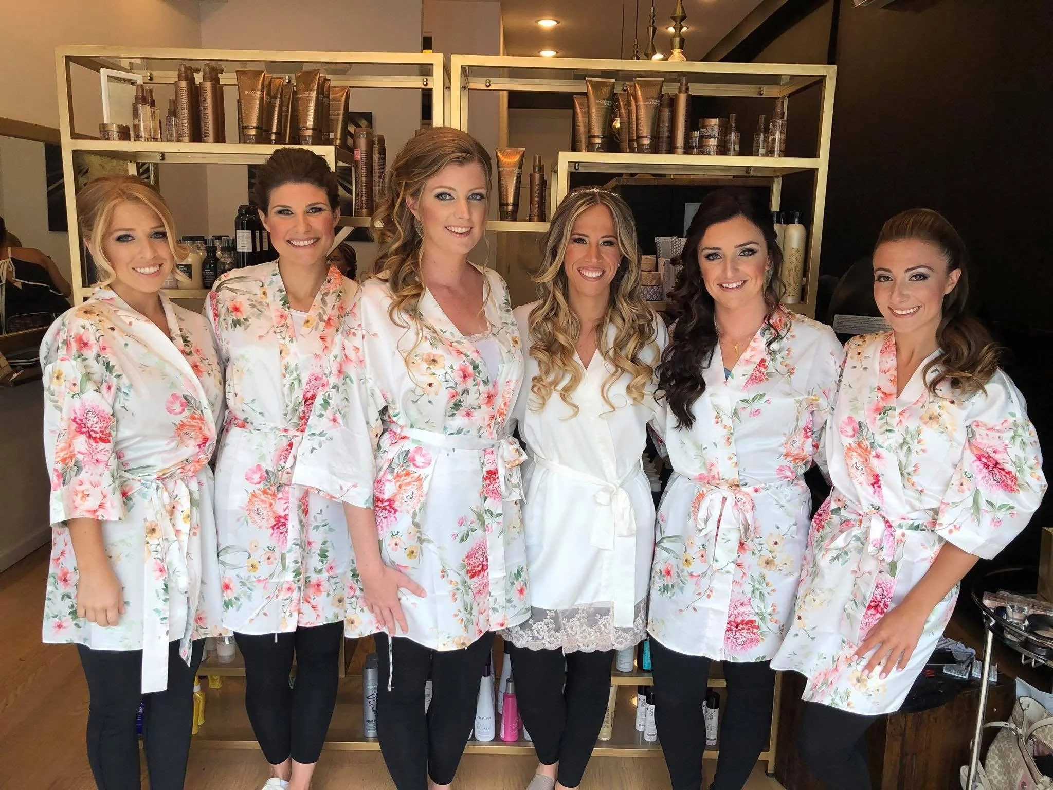 Six women standing in a row wearing matching floral robes, smiling, with shelves of hair products behind them in a salon or spa setting.