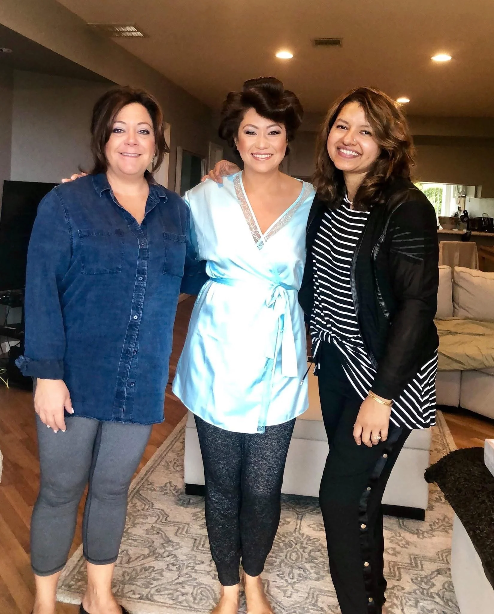 Three women standing together in a living room, smiling at the camera. The woman in the middle is wearing a light-colored satin robe, and the other two women are dressed casually in dark-colored clothing. They are close to each other, with arms aroun