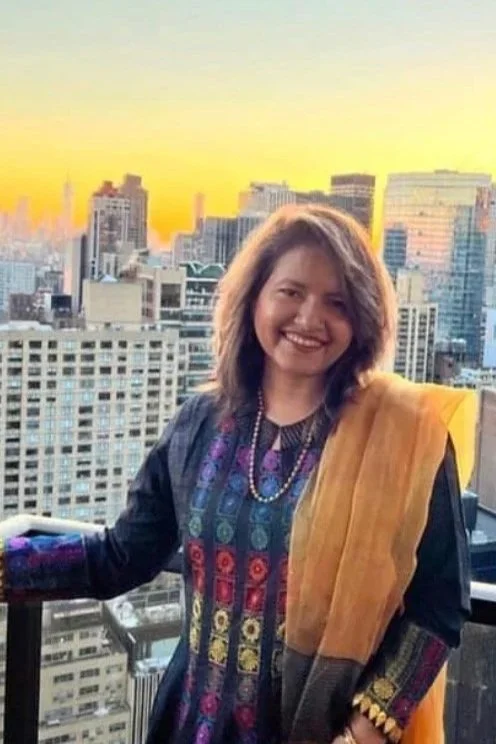 A woman smiling on a rooftop balcony with a city skyline at sunset in the background. She is wearing a colorful dress and a mustard-colored shawl.