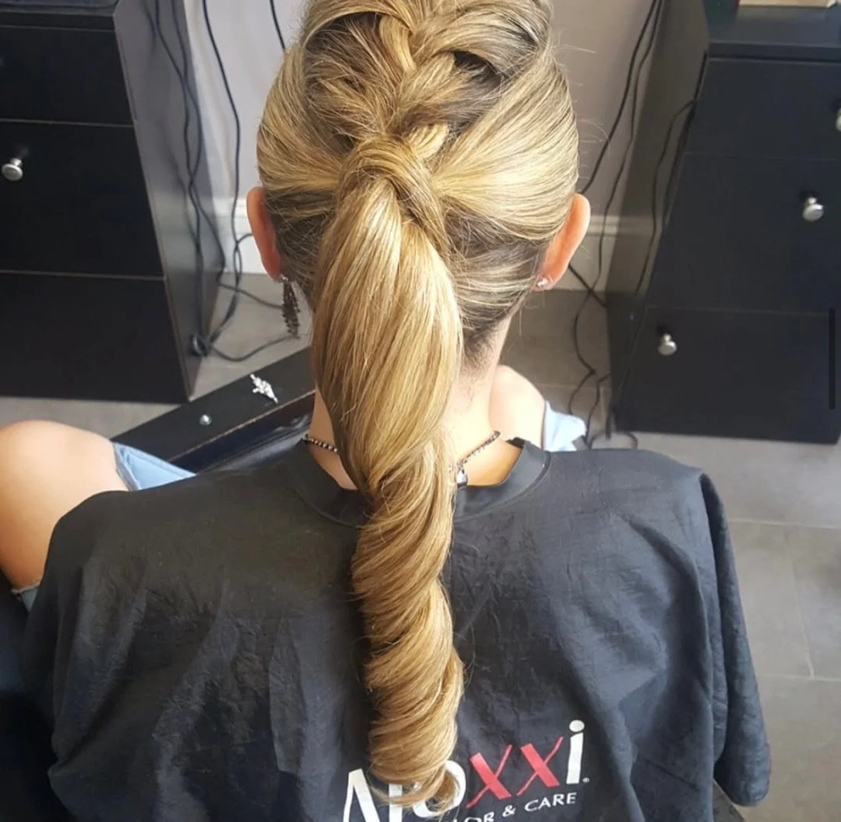 Back view of a woman with long blonde hair styled in a twisted and curled ponytail at a hair salon.