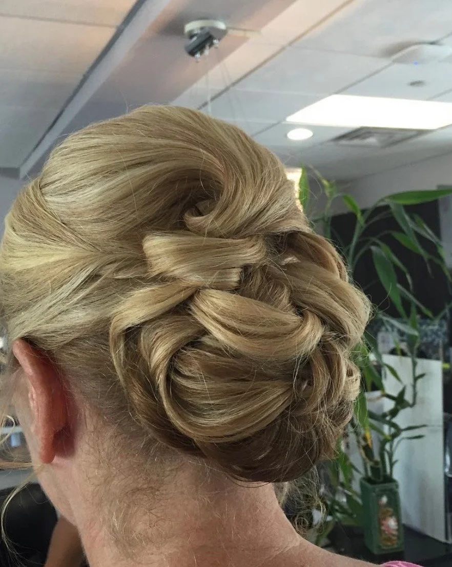 Elegant blonde woman's updo hairstyle with twists and curls, set in an indoor space with ceiling lights and green plants in the background.