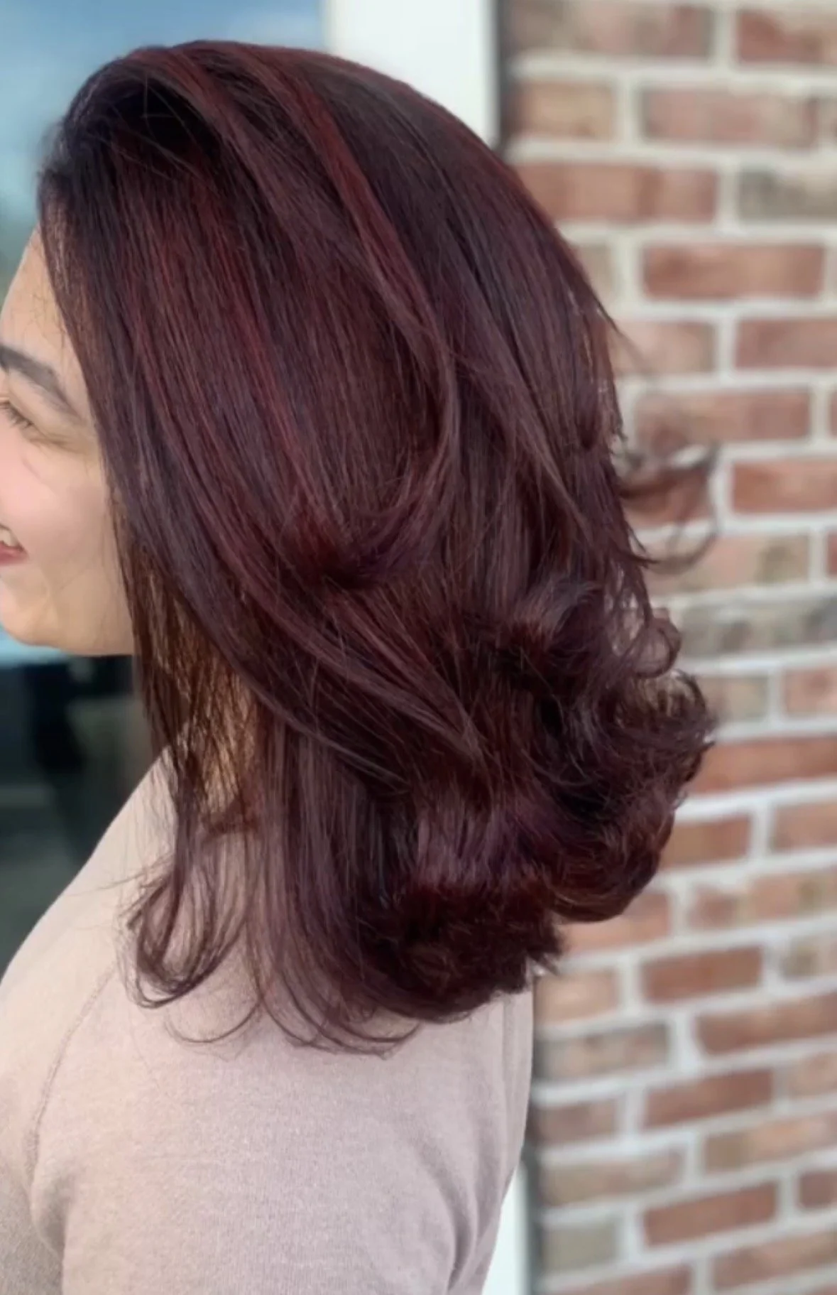 Side view of a woman with medium-length, burgundy styled hair, standing in front of a brick wall.