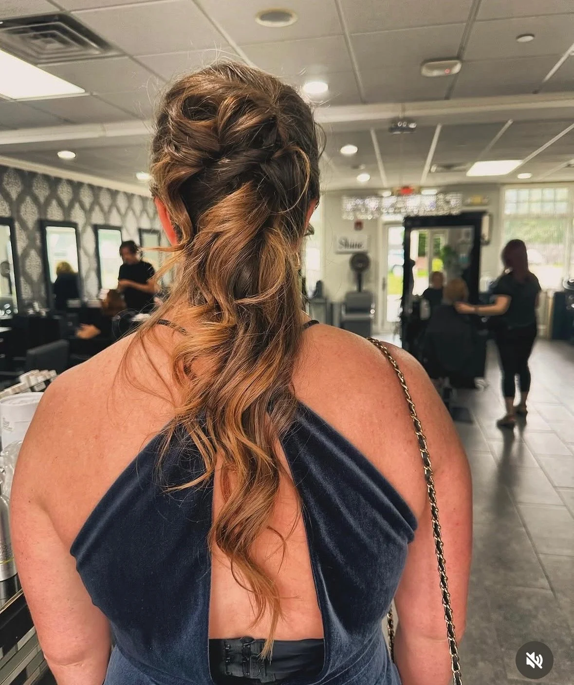 A woman with styled half-up, half-down curly hair in a salon, wearing a dark sleeveless top with a chain strap, facing away from the camera, in a busy salon setting with other clients and staff in the background.