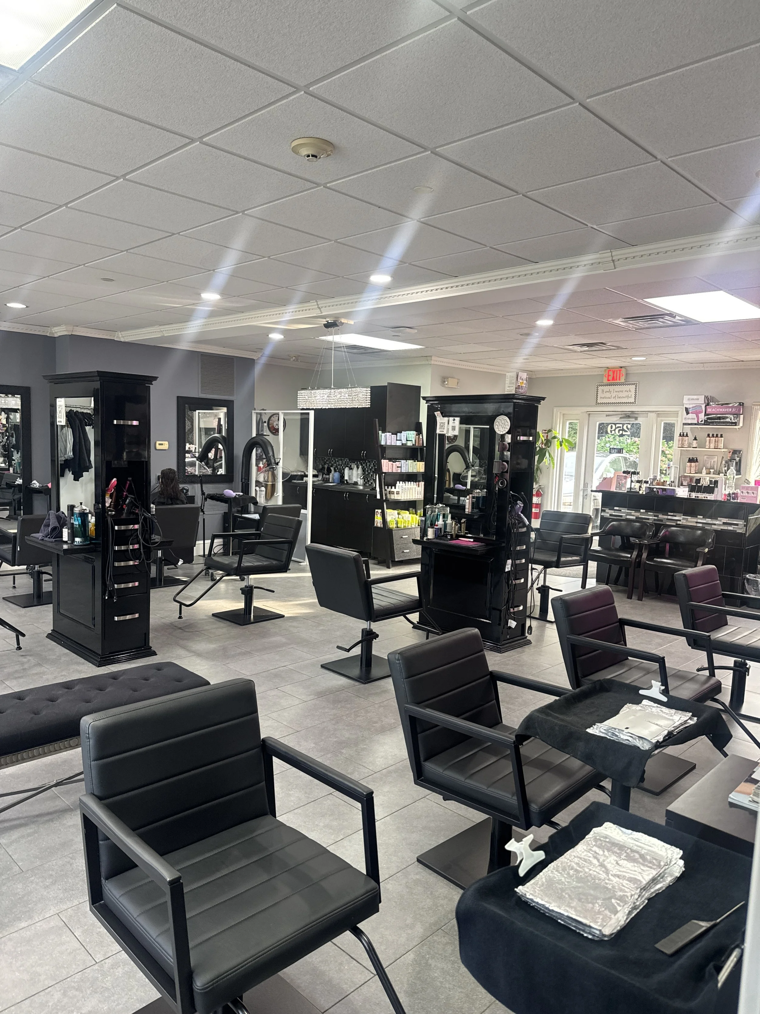 Interior of a hair salon with black styling chairs, mirrors, and salon products on shelves. Bright, evenly lit with a tiled ceiling and large windows at the back.