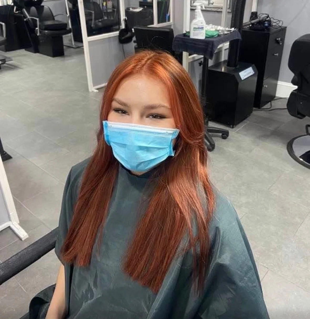 A woman with long red hair and a blue face mask sitting in a hair salon chair.