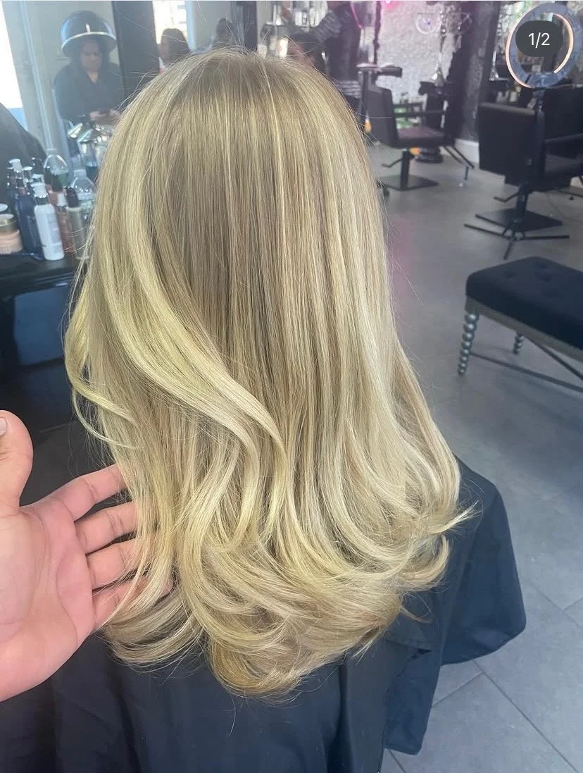 Back view of a woman with long, wavy blonde hair sitting in a salon chair.