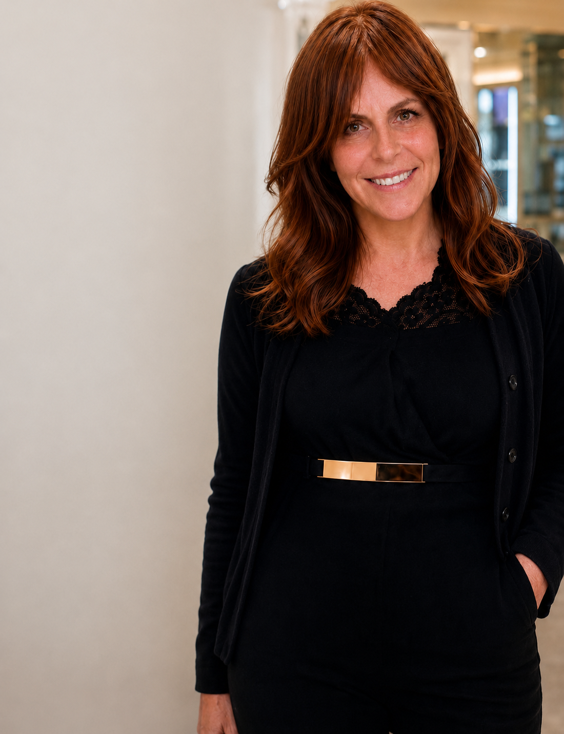 A middle-aged woman with shoulder-length red hair smiling, wearing a black top with lace detailing, a black cardigan, and a metallic belt, standing indoors in front of a plain wall.