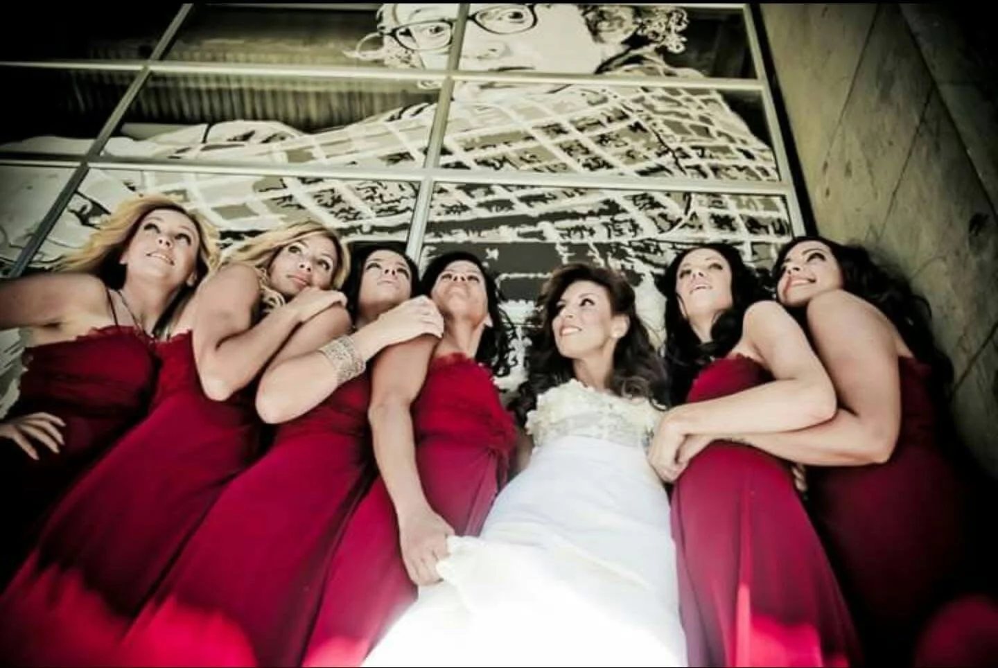 A group of women dressed in formal dresses, lying on their sides in a circle and looking upward, with an industrial-style ceiling with artwork above them.