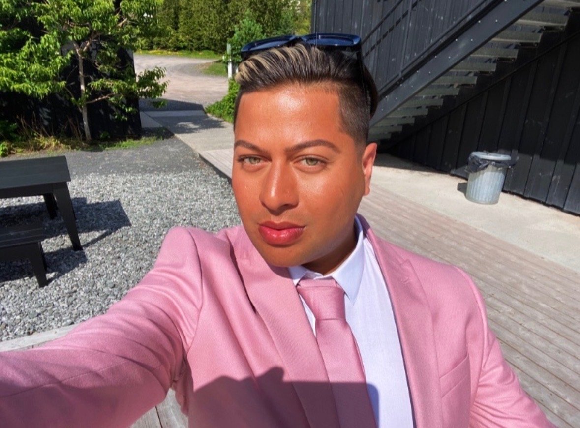 A person taking a selfie outdoors, wearing a pink suit, white shirt, and pink tie, with sunglasses resting on their head.