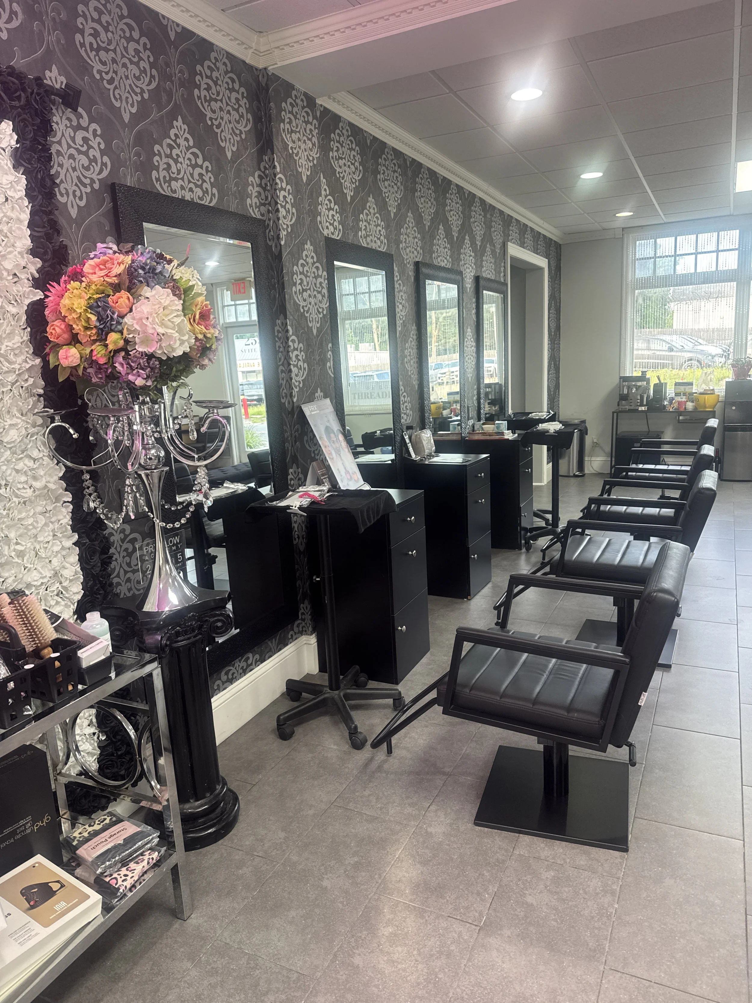 A hair salon interior with black styling chairs arranged in a row facing mirrors on a black damask-patterned wall. There is a cart with hair styling tools and products, and a floral centerpiece with purple, pink, and white flowers on a tall silver stand. Large windows at the front let in natural light, revealing parked cars outside.