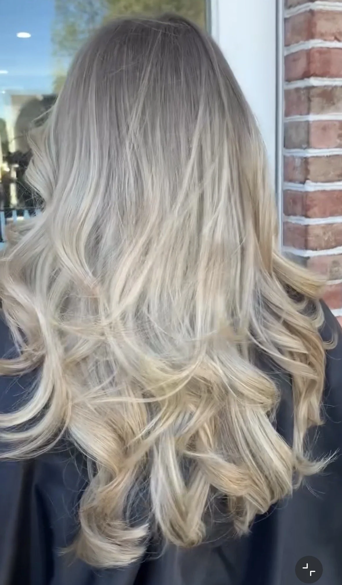 Back of a person with long, blonde, styled hair with waves and curls, standing near a window with a brick wall.