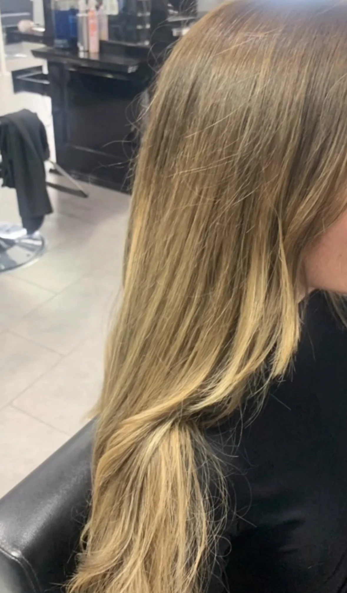 Close-up of a woman with long, blonde hair sitting in a salon or barbershop chair, with styling tools and products visible in the background.