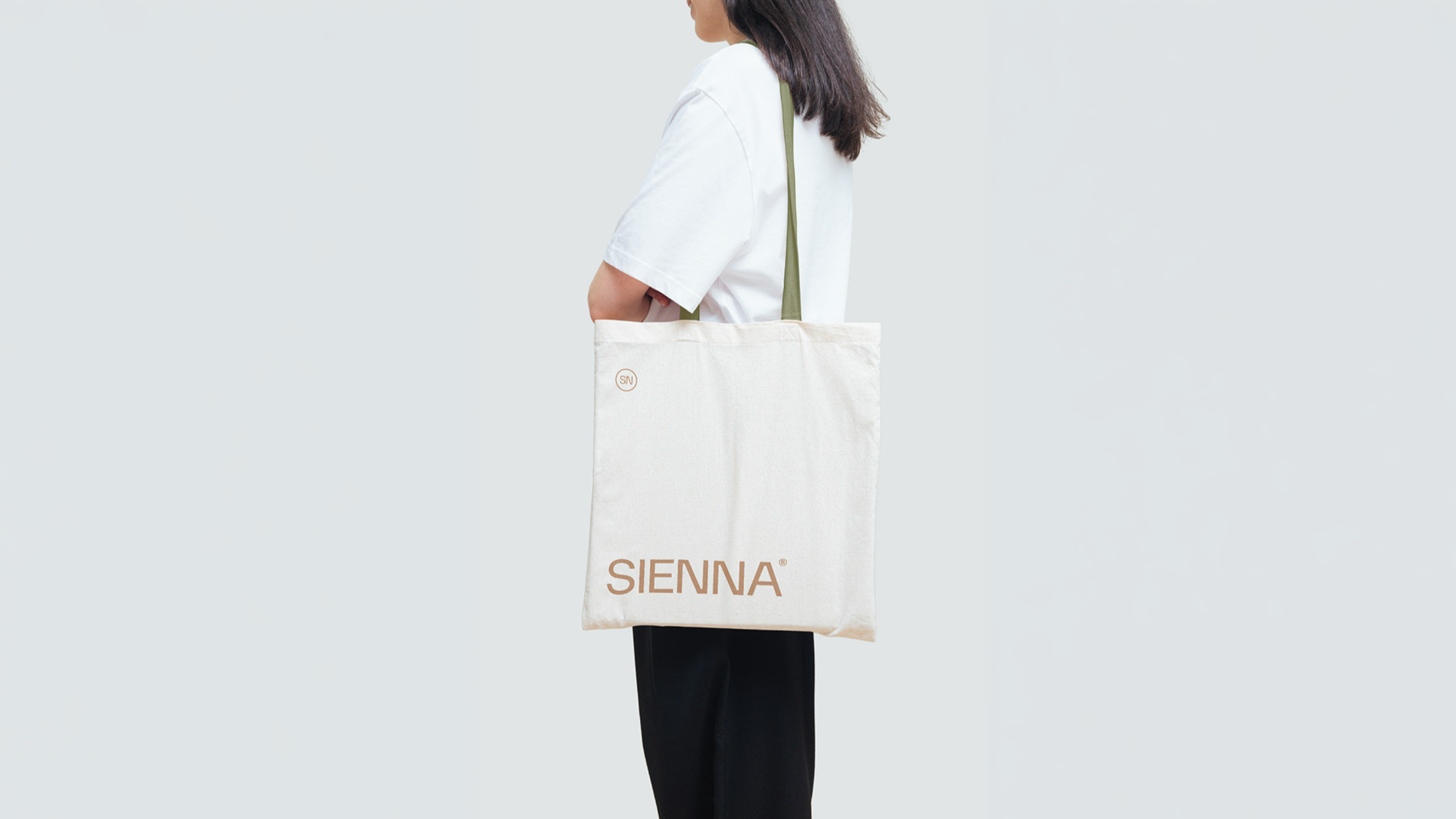 Mujer con camiseta blanca y bolso de tela con las palabras SIENNA en letras grandes, de perfil, de espaldas en un fondo gris claro.