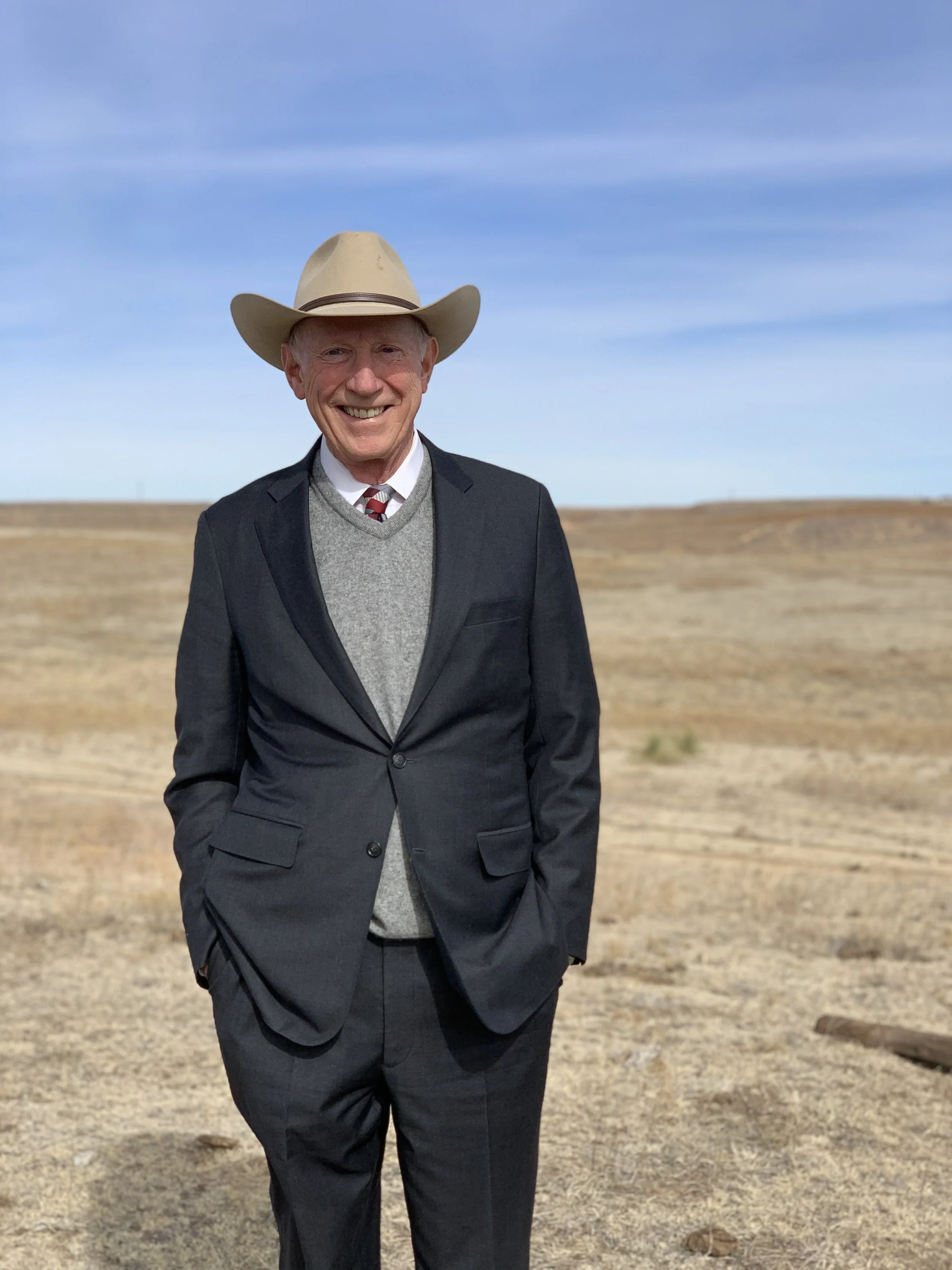 An elderly man in a suit with a red and white striped tie, gray sweater, and black coat is smiling while walking outdoors in a barren field under a blue sky. He is wearing a wide-brimmed beige hat.