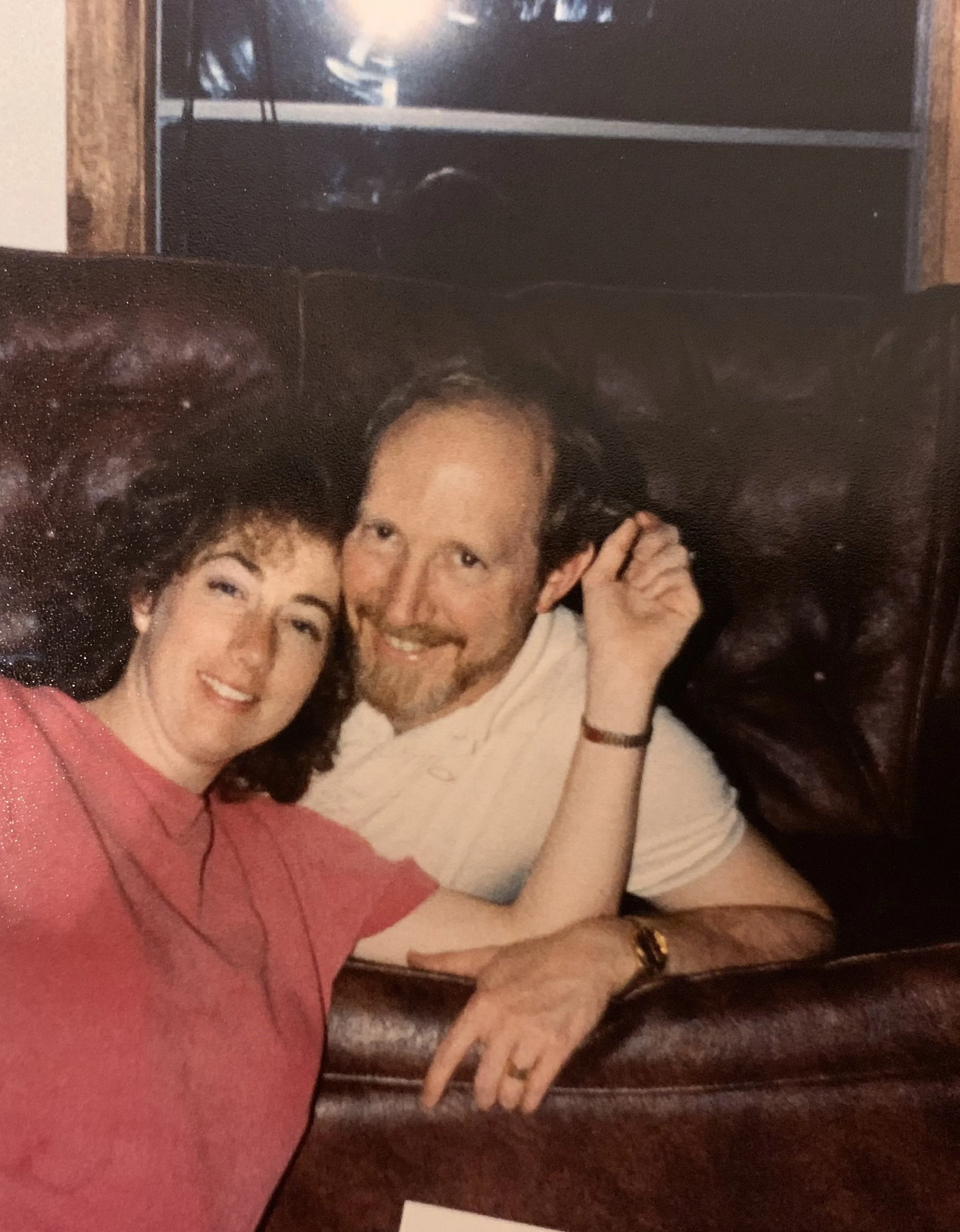 A woman with curly dark hair and a man with straight brown hair sitting closely together in a restaurant booth. The woman is wearing a red top, and the man is wearing a white shirt. They are smiling and seem to be happy.
