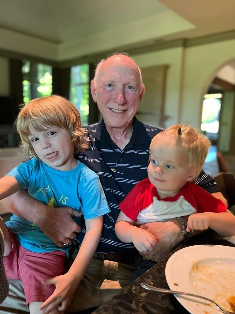 An elderly man smiling and holding two young children, one on each side, in a restaurant setting with large windows and a high ceiling.