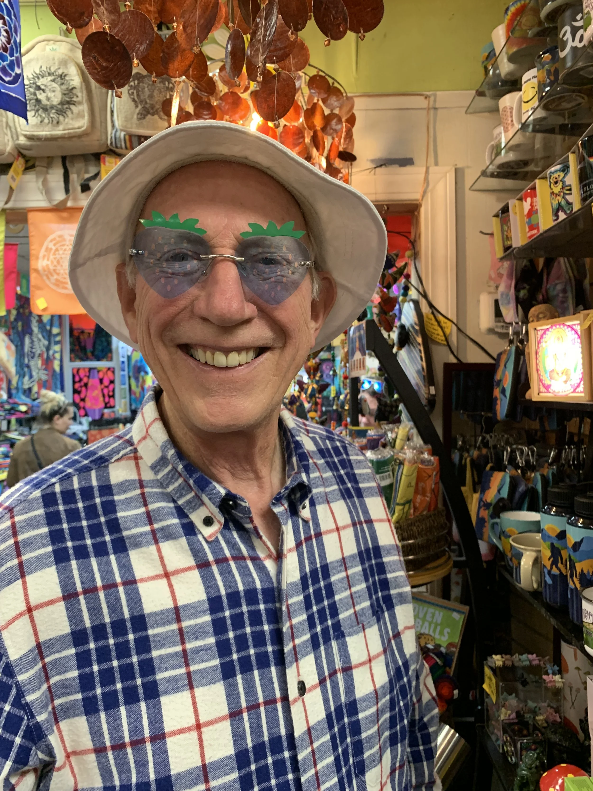 A smiling man wearing novelty pineapple-shaped sunglasses and a white bucket hat, standing inside a colorful gift shop with various items, including mugs, posters, and decorations, visible in the background.