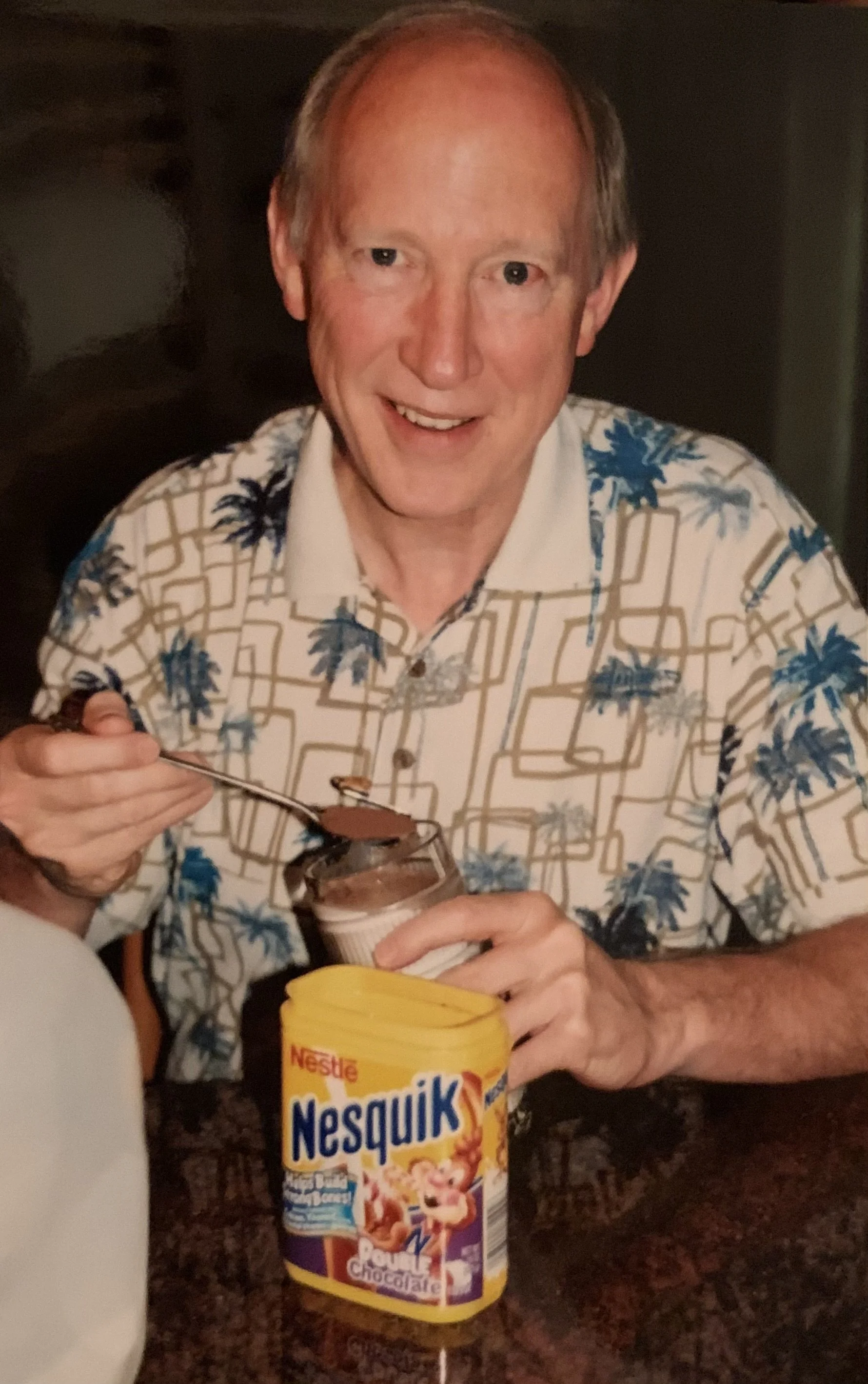 A man in a Hawaiian-style shirt is holding a spoonful of Nesquik chocolate powder over a container of the same brand, in a kitchen setting.