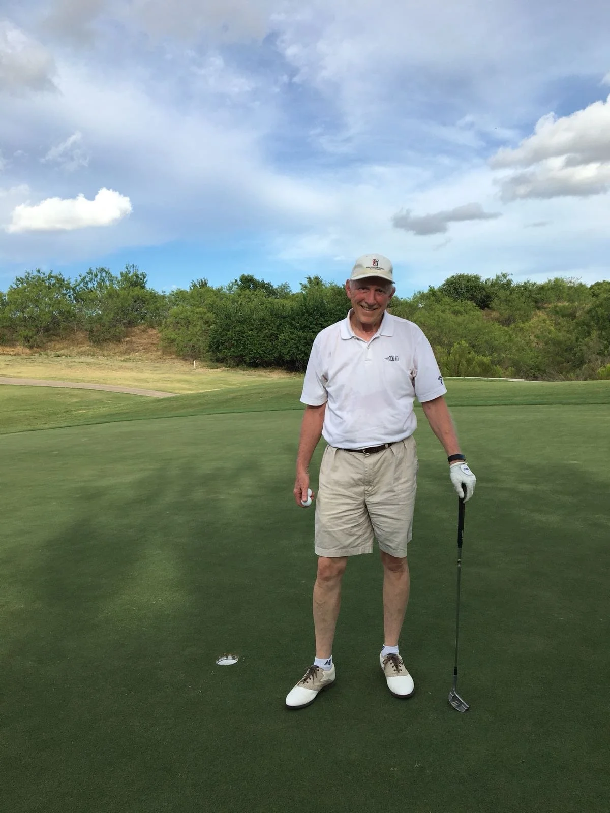 An elderly man smiling on a golf course holding a golf club and ball, wearing a white polo shirt, beige shorts, a white cap, and golf shoes, with trees and a cloudy sky in the background.