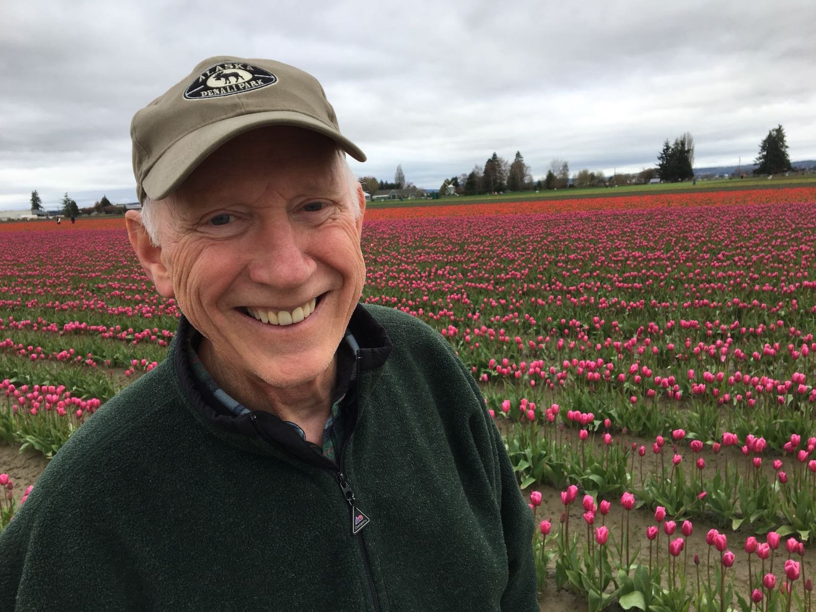 A smiling man wearing a beige cap with a park logo, standing in a vast field of pink tulips under a cloudy sky.