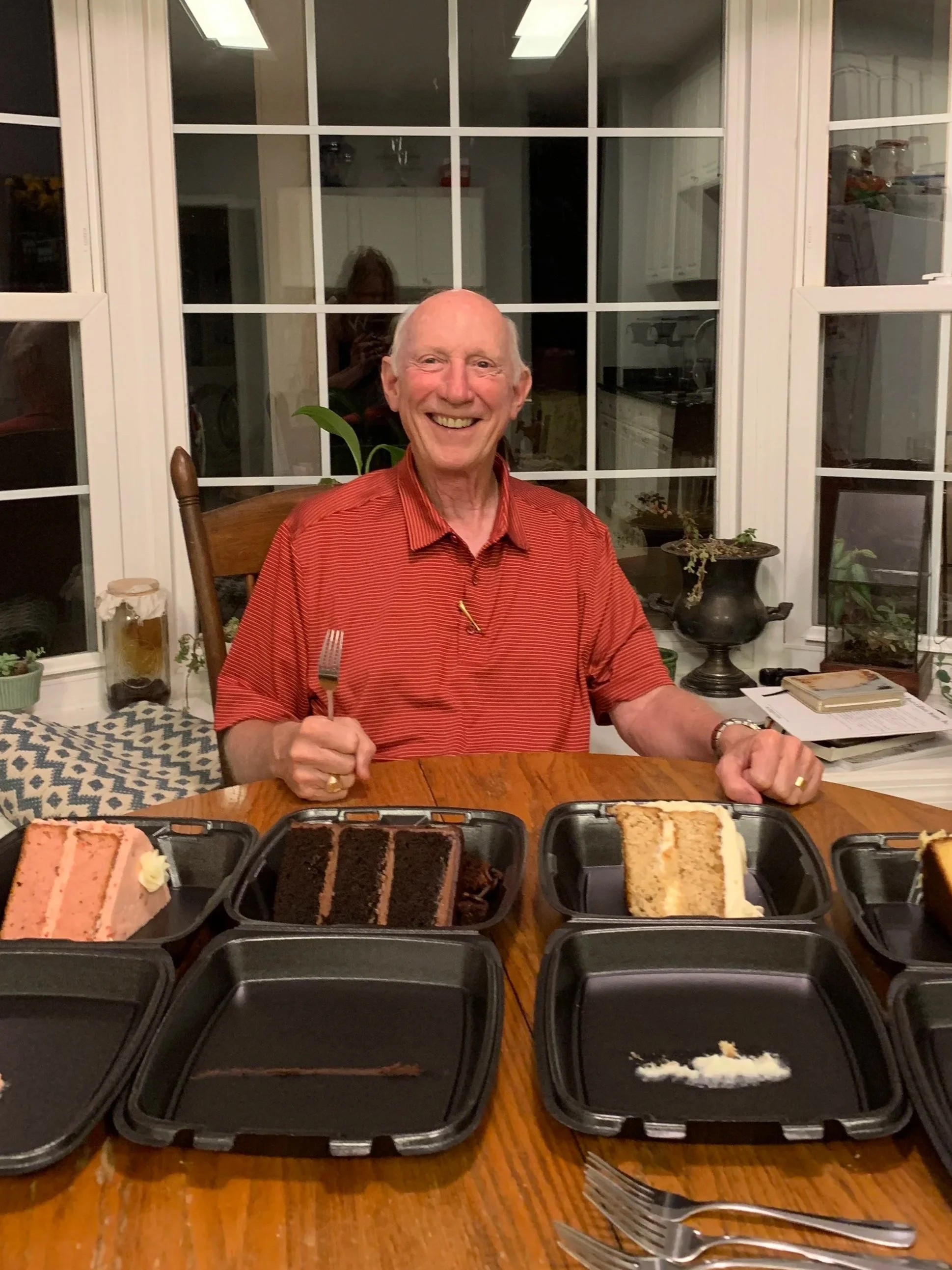 A smiling elderly man in a red striped shirt sitting at a wooden table with slices of cake in black takeout containers in front of him. The background shows a large window with white framing and a kitchen. There are utensils and a notebook on the table.