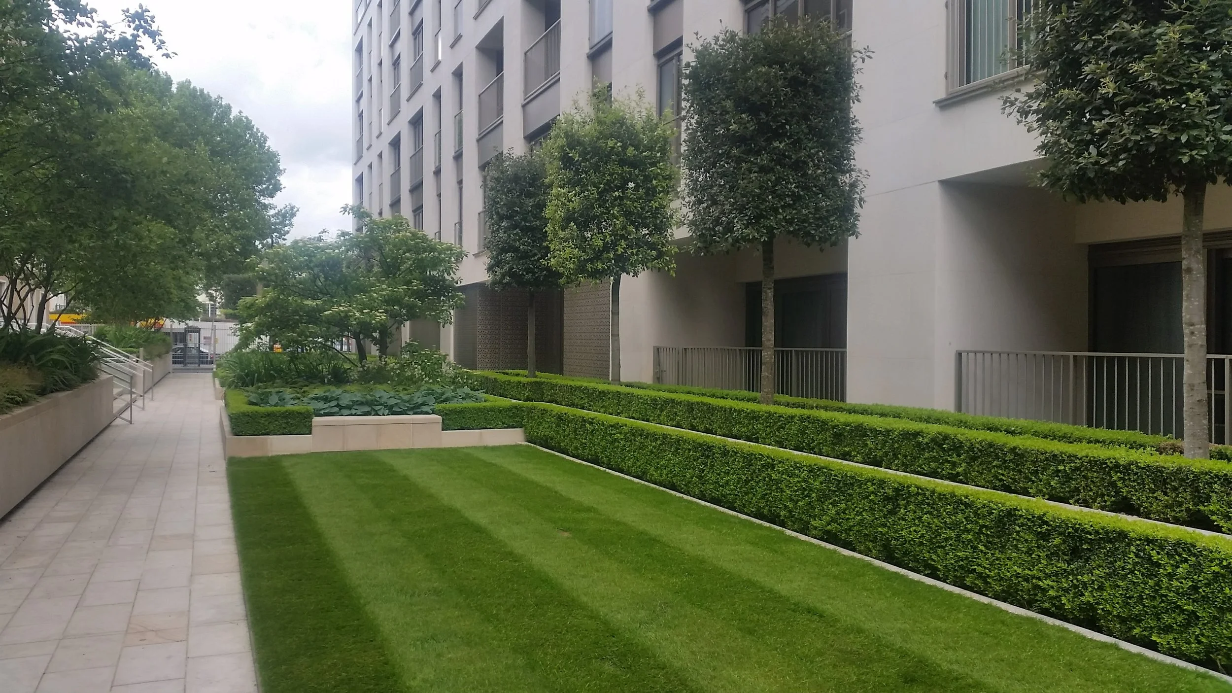 Modern residential building with landscaped garden, trimmed bushes, and trees along a paved walkway.