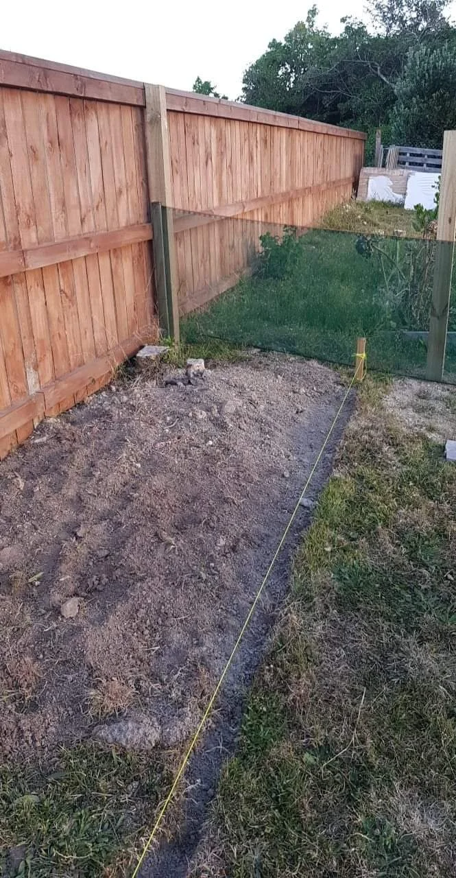 A backyard with a newly dug strip of dirt and a yellow string line, with a wooden fence on the left and some greenery on the right, indicating yard renovation or landscaping in progress.