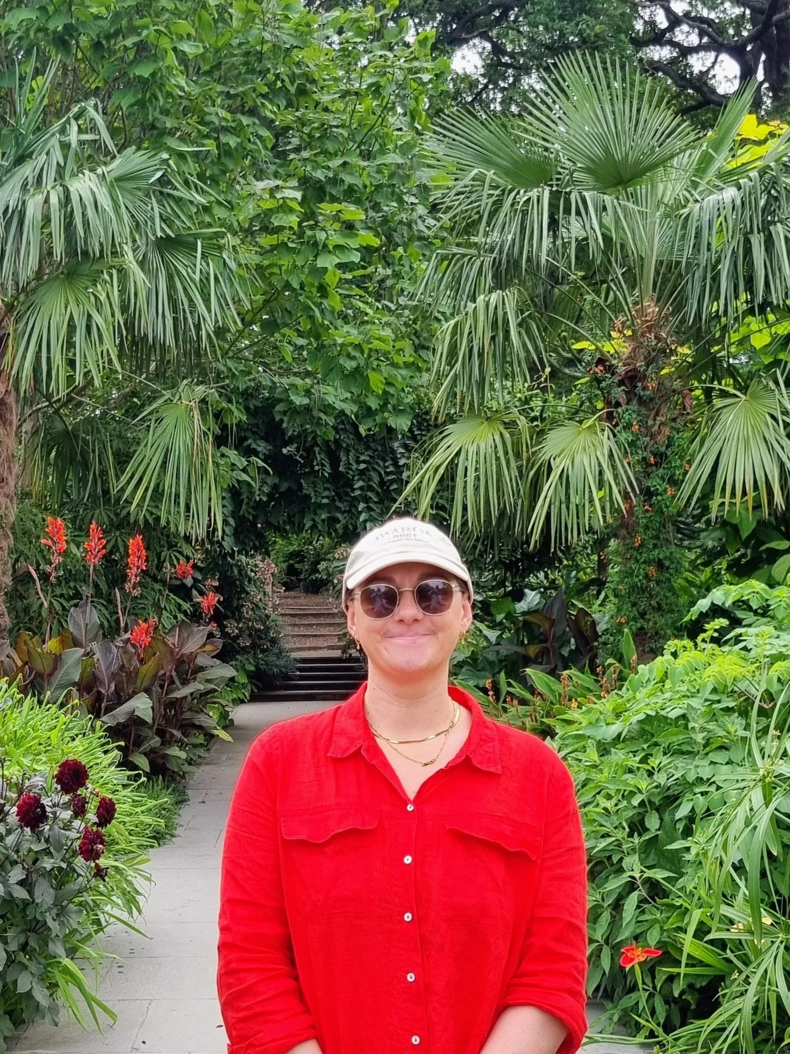A woman in a red shirt and sunglasses standing in a lush, green garden with a staircase behind her.