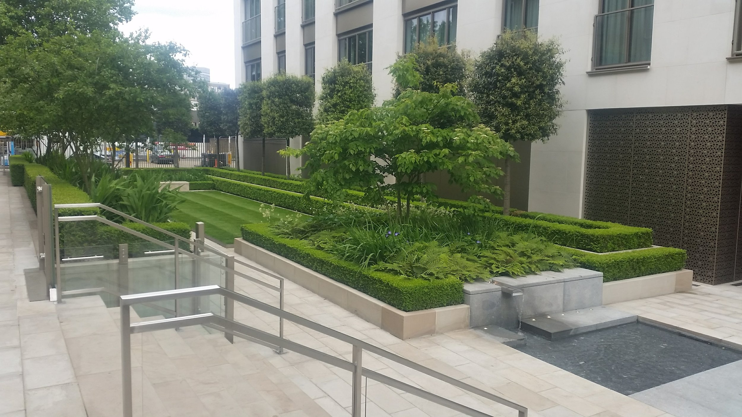 Urban courtyard with landscaped garden, trimmed bushes, small trees, and a building with large windows and decorative metal panels.