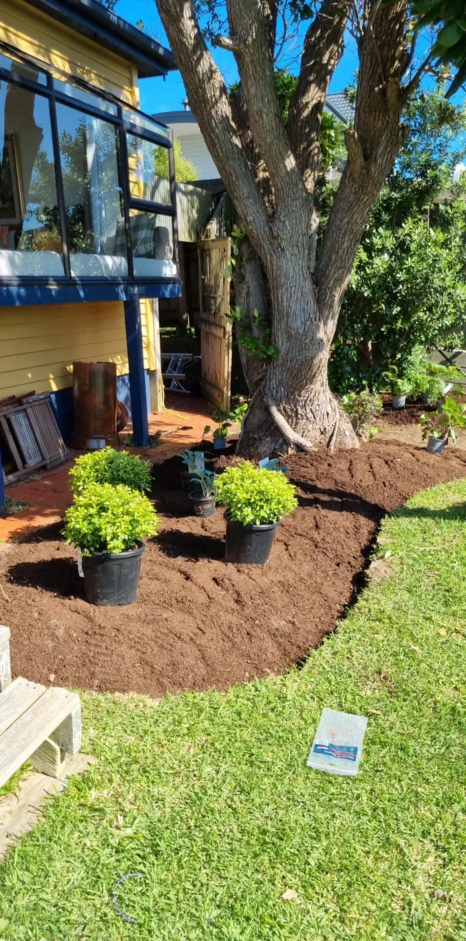 A garden area adjacent to a yellow house features a lawn with newly tilled soil, three potted plants, and a large tree with textured bark. Items like a watering can and gardening supplies are visible, along with outdoor furniture and a screened porch.
