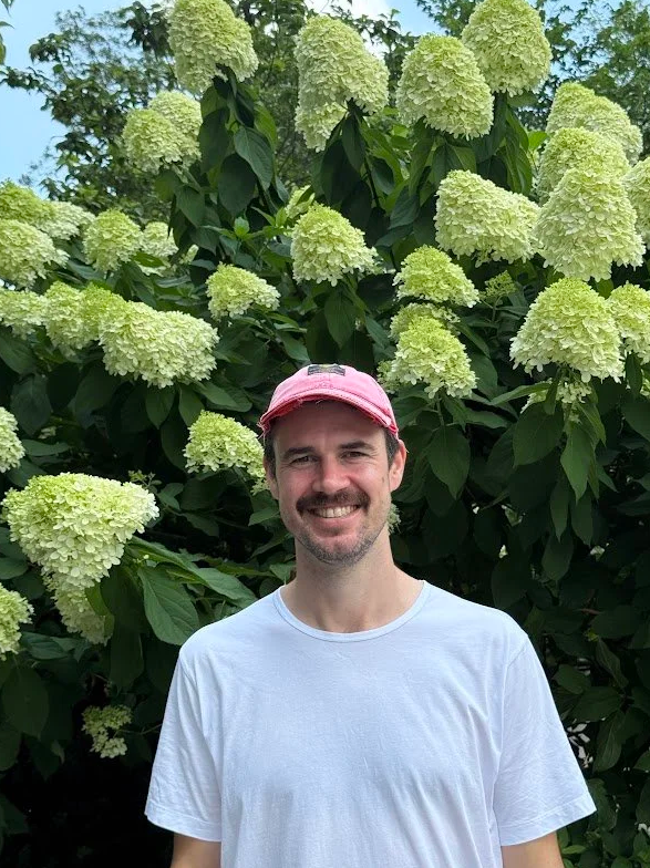 A man smiling and wearing a pink cap and white t-shirt, standing in front of large, blooming white hydrangea flowers and green foliage.