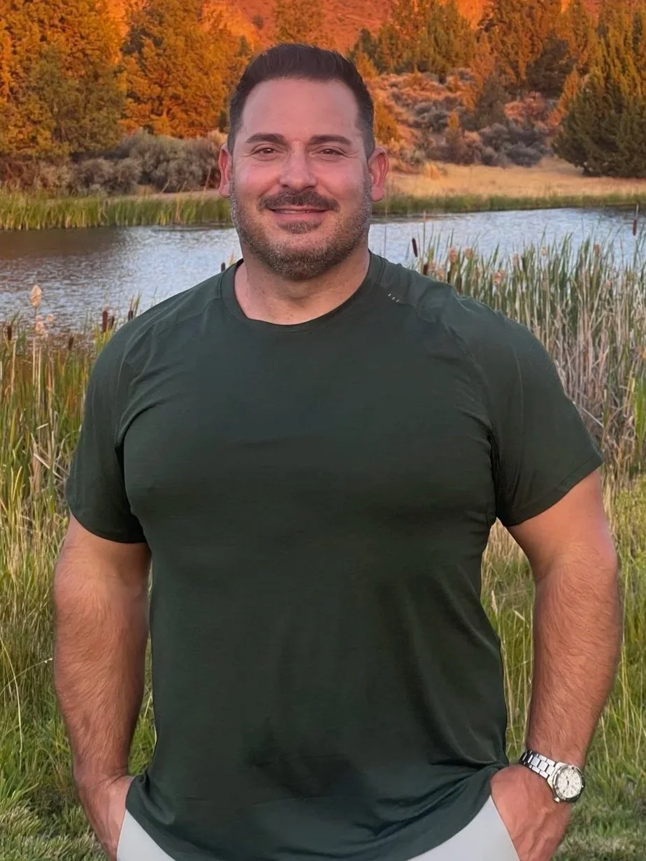 A man standing outdoors near a lake with tall grass and trees, during sunset, smiling, wearing a dark green t-shirt and a silver watch.