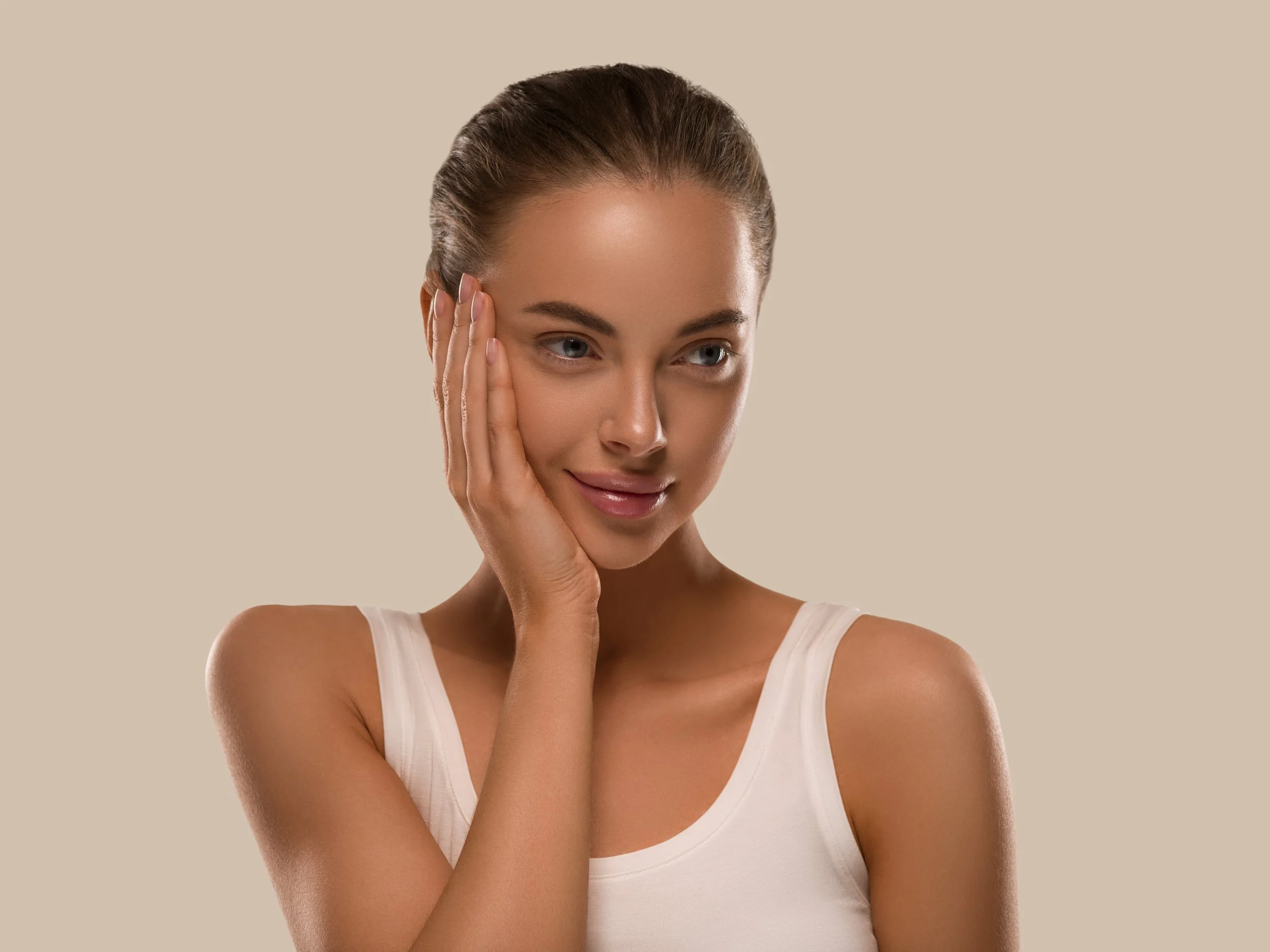 A young woman with brown hair styled in a slicked-back bun, wearing a white tank top, gently touching her face with a serene expression, against a beige background.